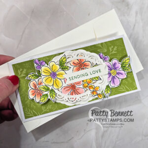 A hand holds a floral greeting card with colorful yellow, purple, and orange flowers on a green background. The card features a white decorative frame and the words "SENDING LOVE" in the center. The envelope and a watermark reading "Patty Bennett PATTYSTAMPS.com" are visible. To read more, visit Patty Stamps (pattystamps.com)
