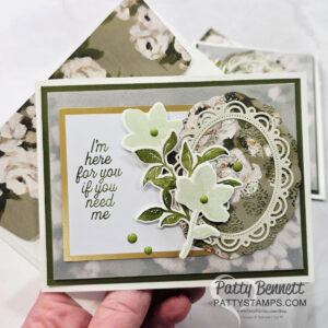 A handmade floral greeting card with layered green and beige papers, featuring die-cut flowers, ornate oval frames, and the message, "I’m here for you if you need me." The background shows a matching floral envelope. Card made by Patty Bennett. To read more, visit Patty Stamps (pattystamps.com)