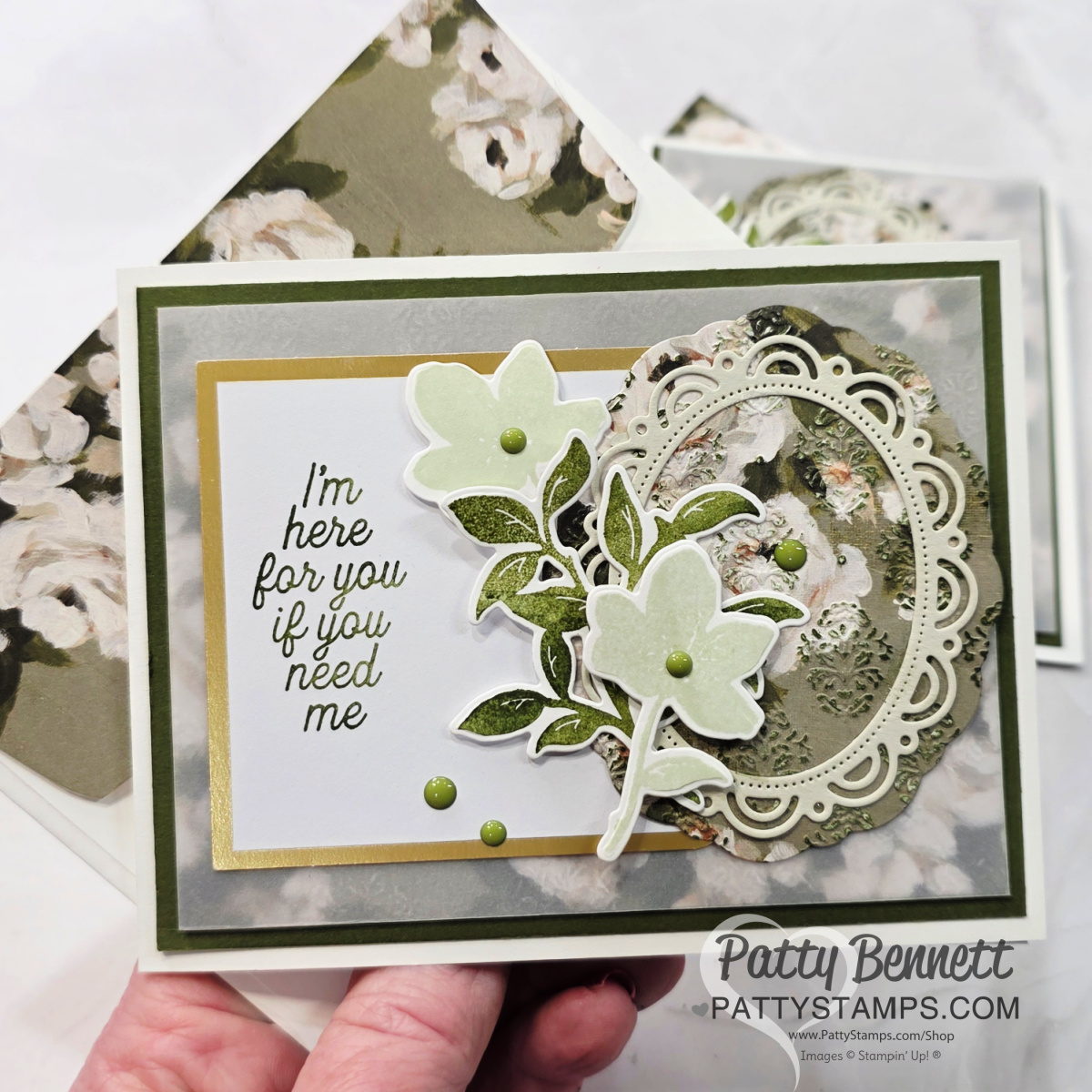 A handmade floral greeting card with layered green and beige papers, featuring die-cut flowers, ornate oval frames, and the message, "I’m here for you if you need me." The background shows a matching floral envelope. Card made by Patty Bennett. To read more, visit Patty Stamps (pattystamps.com)