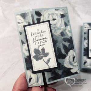 A hand holds a floral greeting card with blue, gray, and white rose patterns. The front features a black flower illustration and a message: "If friends were flowers, I'd pick you." The card edges are a pale blue-gray. The background is softly blurred. To read more, visit Patty Stamps (pattystamps.com)