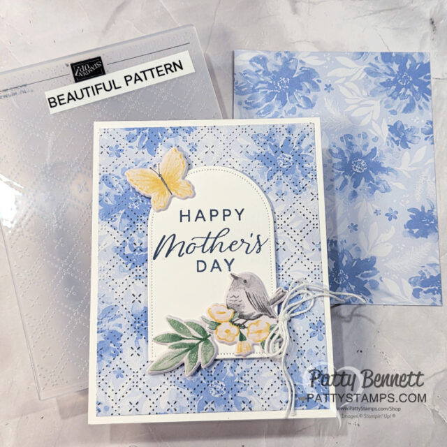 Embossing your designer paper. A handmade Mother’s Day card with blue floral patterns, a yellow butterfly, and a small bird cutout. The card says “Happy Mother’s Day” in blue text. An embossed patterned folder and a floral envelope are also shown in the background. To read more, visit Patty Stamps (pattystamps.com)