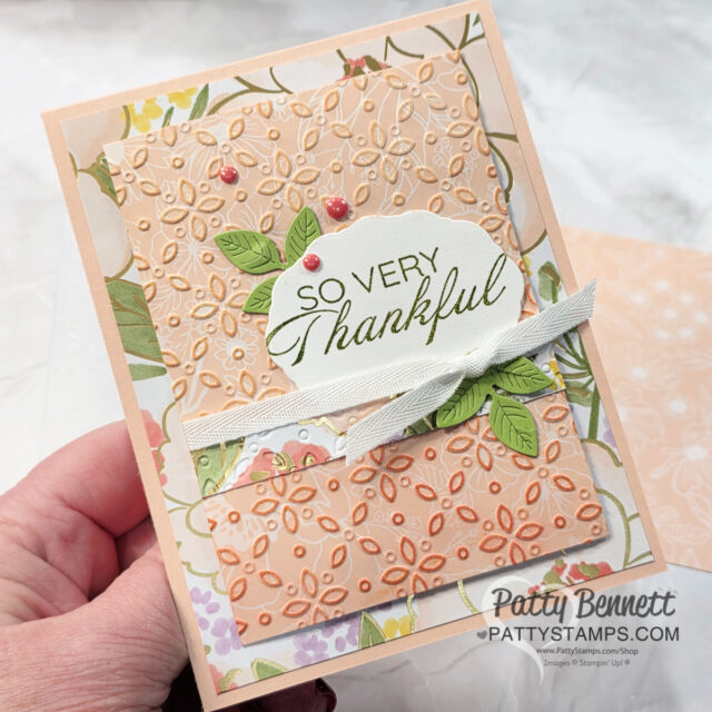 A hand holds a handmade card decorated with peach and green floral patterns, embossed flowers, a white ribbon, green leaves, and the message "SO VERY Thankful" in elegant script. Pink pearls embellish the card. The Patty Stamps logo is visible. To read more, visit Patty Stamps (pattystamps.com)