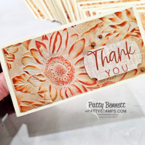 A hand holds a handmade thank you card featuring an embossed sunflower design in shades of orange and beige. “Thank You” is written on textured paper, accented by two small brown dots. Several similar cards are stacked in the background. To read more, visit Patty Stamps (pattystamps.com)