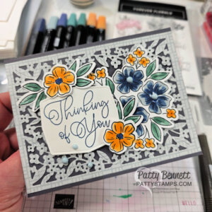 A handmade greeting card with intricate gray and silver floral patterns, accented by bright orange and blue flower embellishments and green leaves. In the center, elegant script reads “Thinking of You.” Three blue enamel dots add decoration. To read more, visit Patty Stamps (pattystamps.com)