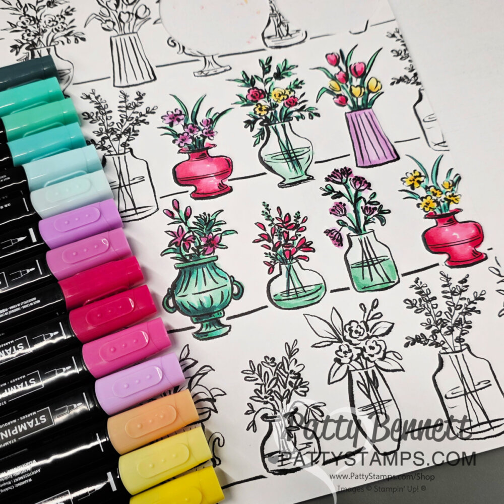 A partially colored page shows black-and-white sketches of flower vases. Some vases and flowers are brightly colored with markers in pink, teal, and yellow shades. Several dual-tip markers in similar colors are arranged to the left of the page. To read more, visit Patty Stamps (pattystamps.com)