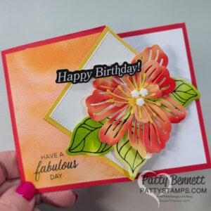 A handmade fun fold birthday card features a large layered orange and yellow paper flower with green leaves on a white and gold diamond. The watercolor wash background in orange tones adds charm. Includes “Happy Birthday!” and watercoloring tips, signed “Patty Bennett.”. To read more, visit Patty Stamps (pattystamps.com)