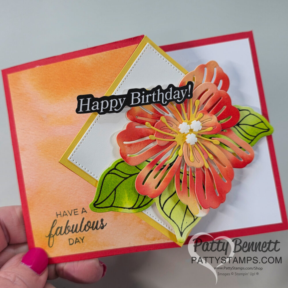 A handmade fun fold birthday card features a large layered orange and yellow paper flower with green leaves on a white and gold diamond. The watercolor wash background in orange tones adds charm. Includes “Happy Birthday!” and watercoloring tips, signed “Patty Bennett.”. To read more, visit Patty Stamps (pattystamps.com)