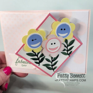 A hand holds a handmade greeting card featuring three smiling cartoon flowers on a layered diamond fun fold background. Green leaves and stems are below, with a soft pink polka dot base and “fabulous day” message—perfect for sharing in your next video!. To read more, visit Patty Stamps (pattystamps.com)
