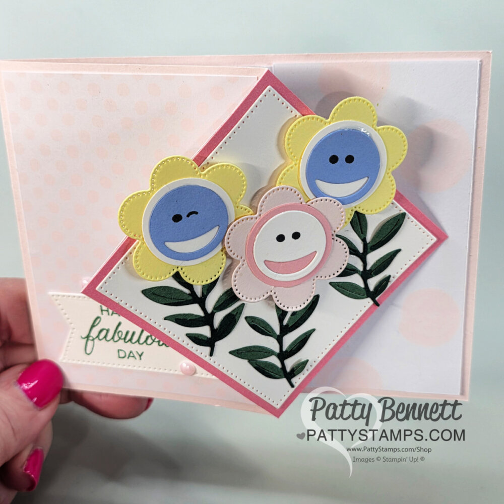 A hand holds a handmade greeting card featuring three smiling cartoon flowers on a layered diamond fun fold background. Green leaves and stems are below, with a soft pink polka dot base and “fabulous day” message—perfect for sharing in your next video!. To read more, visit Patty Stamps (pattystamps.com)