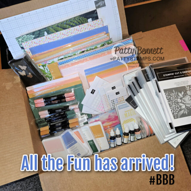 An open cardboard box filled with colorful scrapbooking and stamping supplies, including decorative paper, ink pads, markers, embossing folders, blending brushes, and bottles of ink. Text on the image reads, “All the Fun has arrived! #BBB”. To read more, visit Patty Stamps (pattystamps.com)