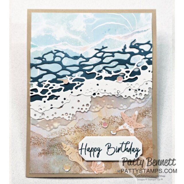 A handmade birthday card with a layered die-cut seashore scene: blue sky, white foamy waves, sandy beach, seashells, and a starfish. The text "Happy Birthday" is on a white label at the bottom. The card background is tan. To read more, visit Patty Stamps (pattystamps.com)