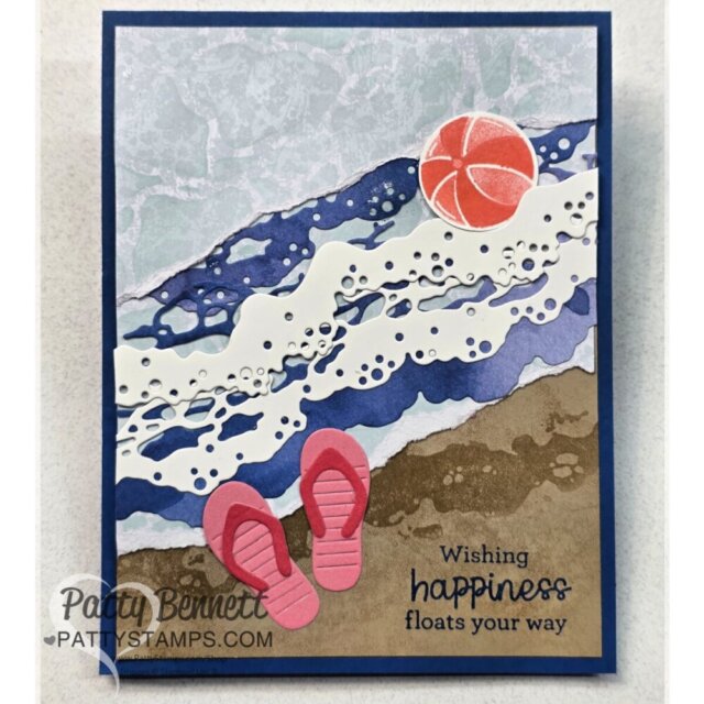 A handmade card shows an ocean scene with layered paper waves, a foamy white border, tan sand, pink flip-flops, and an orange beach ball. The card reads, "Wishing happiness floats your way." The PattyStamps.com watermark is in the lower left. To read more, visit Patty Stamps (pattystamps.com)