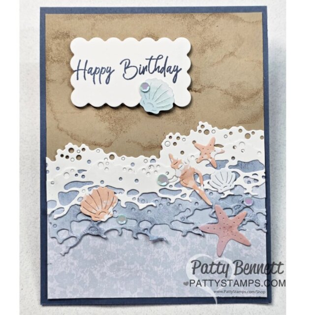 A handmade birthday card features "Happy Birthday" in blue script on a white scalloped panel with a pastel shell. Below, layered die-cut waves and shorelines in beige, white, and blue include pastel seashells and starfishes. The card is signed "Patty Bennett. To read more, visit Patty Stamps (pattystamps.com)