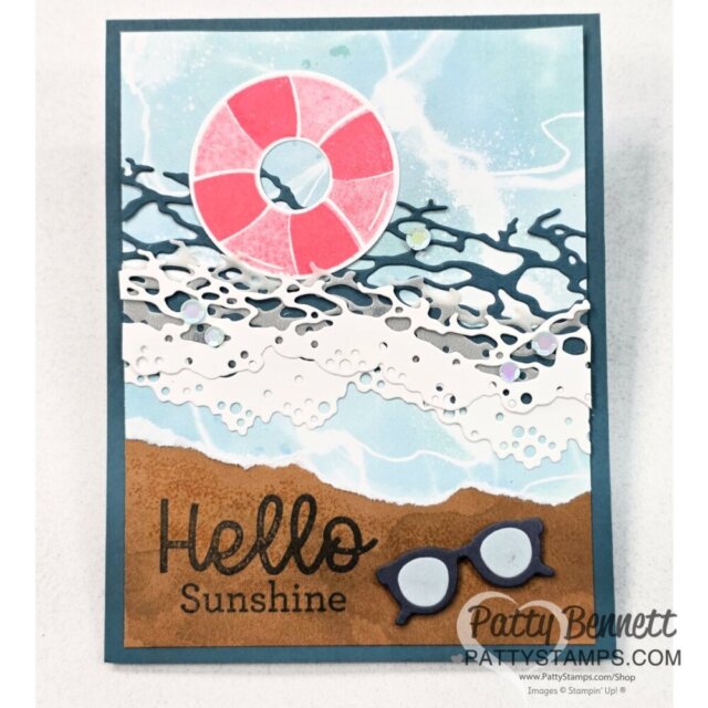 Handmade card with layered paper art: sandy brown beach at the bottom, "Hello Sunshine" text, blue sunglasses, foamy white ocean waves, blue water, a pink and white life preserver, and dark blue coral shapes. Background is light blue sky with clouds. To read more, visit Patty Stamps (pattystamps.com)