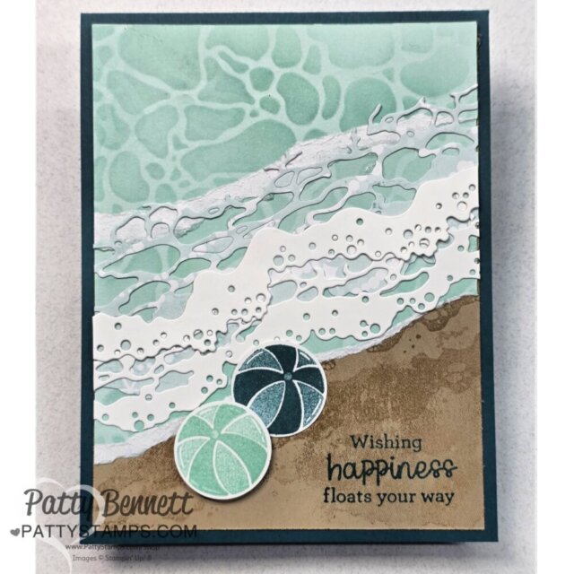 A handmade beach-themed card features textured blue and white waves, a sandy shoreline, and three pastel beach balls. The sentiment at the bottom right reads, “Wishing happiness floats your way.” The artist’s watermark says, "Patty Bennett @PATTYSTAMPS.COM". To read more, visit Patty Stamps (pattystamps.com)