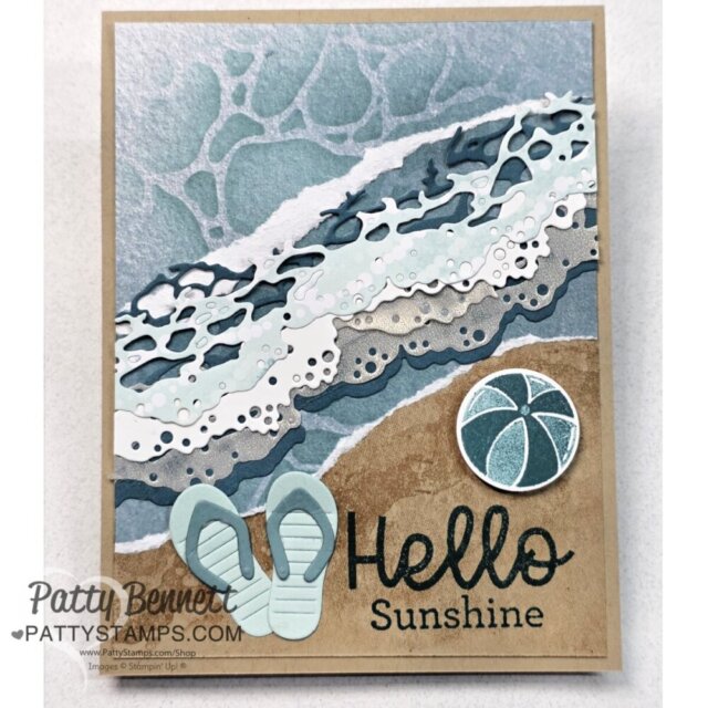 A handmade card shows layered paper art of ocean waves meeting sandy beach. On the sand are a pair of blue flip-flops, a ball, and the words “Hello Sunshine.” The artist’s name, Patty Bennett, appears in the lower left corner. To read more, visit Patty Stamps (pattystamps.com)