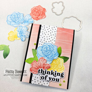 A handmade greeting card features layered die-cut flowers in blue, red, and yellow on a pink and white striped background with a black and white polka dot strip. The card reads "thinking of you" in bold black letters. Crafting supplies are arranged nearby. To read more, visit Patty Stamps (pattystamps.com)