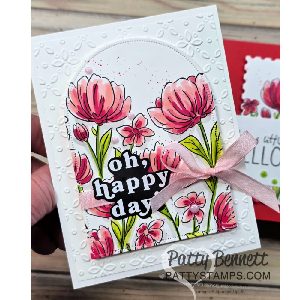 A handmade greeting card features pink flowers with green stems, “oh, happy day” in bold white text on black, and a pink ribbon bow. The card has an embossed white border with a floral pattern and is held in someone’s hand. “Patty Bennett pattystamps.com” is visible. To read more, visit Patty Stamps (pattystamps.com)