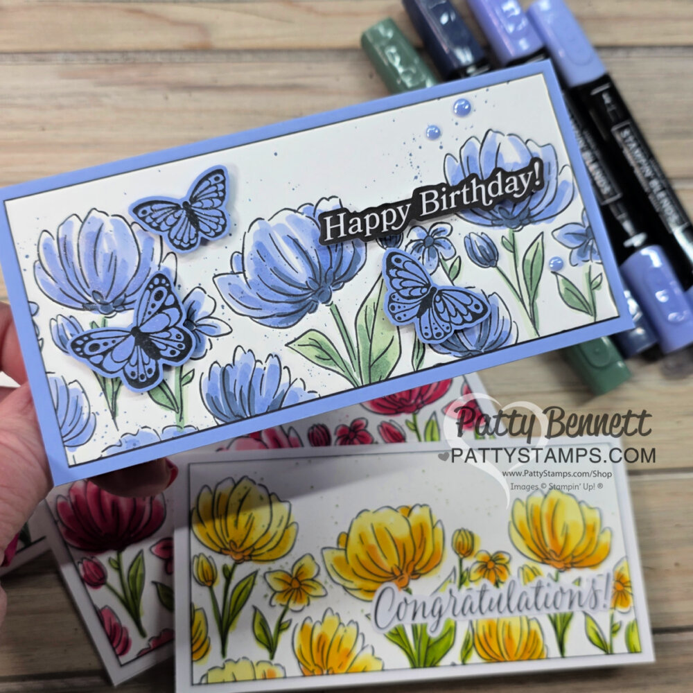 A hand holds a rectangular card decorated with blue and purple flowers, green leaves, and butterflies. “Happy Birthday!” is written in white on a dark banner. Another card below features yellow flowers and “Congratulations!” text. Colored markers are nearby. To read more, visit Patty Stamps (pattystamps.com)