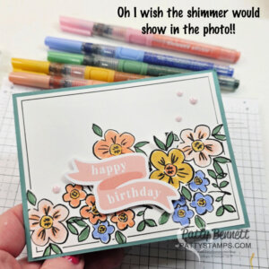 A handmade birthday card is held above a desk with markers. The card features colorful illustrated flowers, a pink banner reading "happy birthday," and three pink embellishments. Text at the top reads, "Oh I wish the shimmer would show in the photo!!. To read more, visit Patty Stamps (pattystamps.com)
