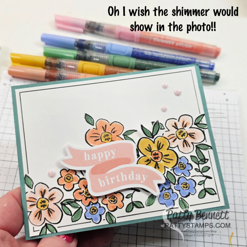 A handmade birthday card is held above a desk with markers. The card features colorful illustrated flowers, a pink banner reading "happy birthday," and three pink embellishments. Text at the top reads, "Oh I wish the shimmer would show in the photo!!. To read more, visit Patty Stamps (pattystamps.com)