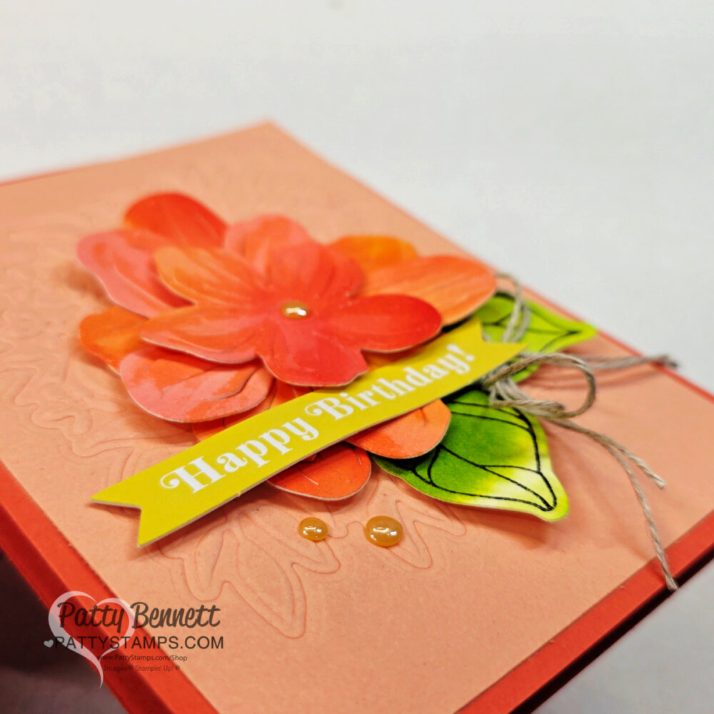 A handmade birthday card features layered orange and red paper flowers, a green and yellow leaf, and a yellow "Happy Birthday!" banner. A twine bow and clear gems accent the design on a peach background. "Patty Bennett" and a watermark appear in the corner. To read more, visit Patty Stamps (pattystamps.com)