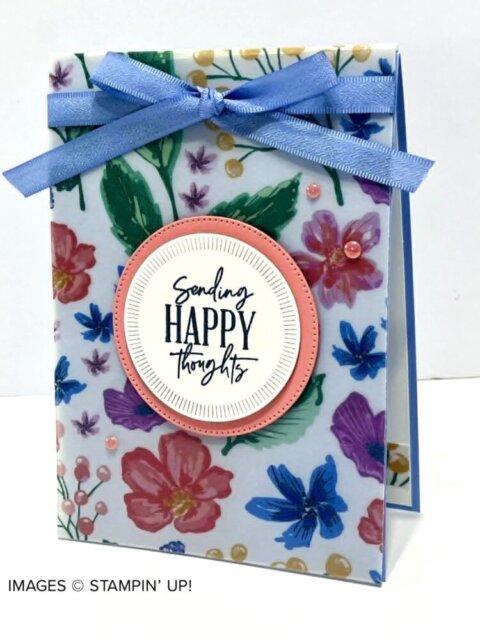 A handmade greeting card features a colorful floral background, topped with a blue ribbon bow. In the center is a round label bordered in pink, with the message “Sending HAPPY thoughts” in bold and script fonts. Pink gems accent the card. To read more, visit Patty Stamps (pattystamps.com)
