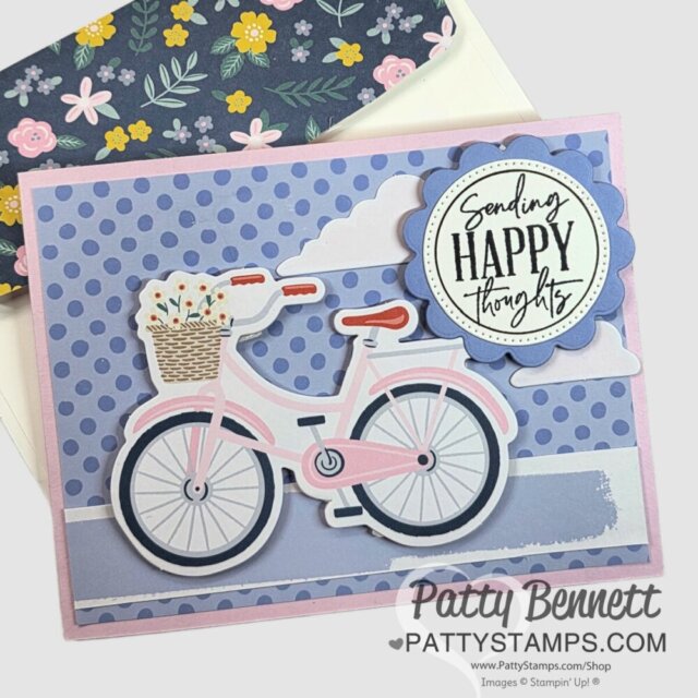 A handmade greeting card features a pink bicycle with a flower basket against a purple polka-dot background. A scalloped blue circle with “Sending HAPPY thoughts” is on the upper right. A floral envelope is partially visible behind the card. To read more, visit Patty Stamps (pattystamps.com)