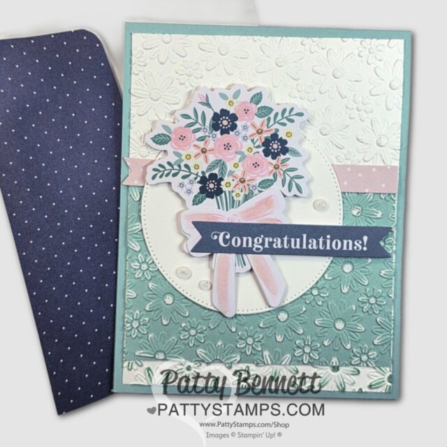 A handmade greeting card features embossed white and mint green floral backgrounds, a bouquet of colorful flowers, a pink ribbon, a “Congratulations!” banner, pearl embellishments, and a matching navy blue envelope with white polka dots. To read more, visit Patty Stamps (pattystamps.com)