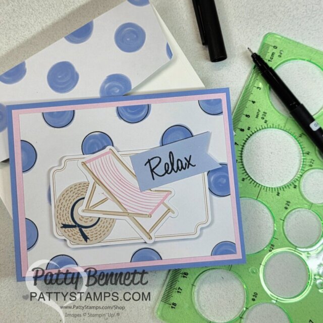 A handmade card with a striped deck chair, sun hat, and a banner reading “Relax” is shown on a blue polka dot background. The card is surrounded by a stencil, black pen, and an envelope with matching blue dots. The PattyStamps watermark is visible. To read more, visit Patty Stamps (pattystamps.com)