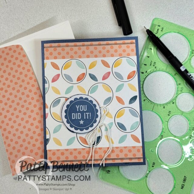 A handmade card with colorful leaf patterns and a “You Did It!” badge is displayed on a desk. Nearby are a green circle template, a black marker, and an orange polka dot envelope. The card is stamped with “Patty Bennett PattyStamps.com.”. To read more, visit Patty Stamps (pattystamps.com)