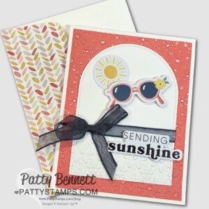 A handmade card features a textured coral border, a white floral-embossed panel, and a black ribbon bow. This quick & easy design is decorated with sun and sunglasses ephemera plus “SENDING sunshine” text. Leaf-pattern envelope beside it. To read more, visit Patty Stamps (pattystamps.com)