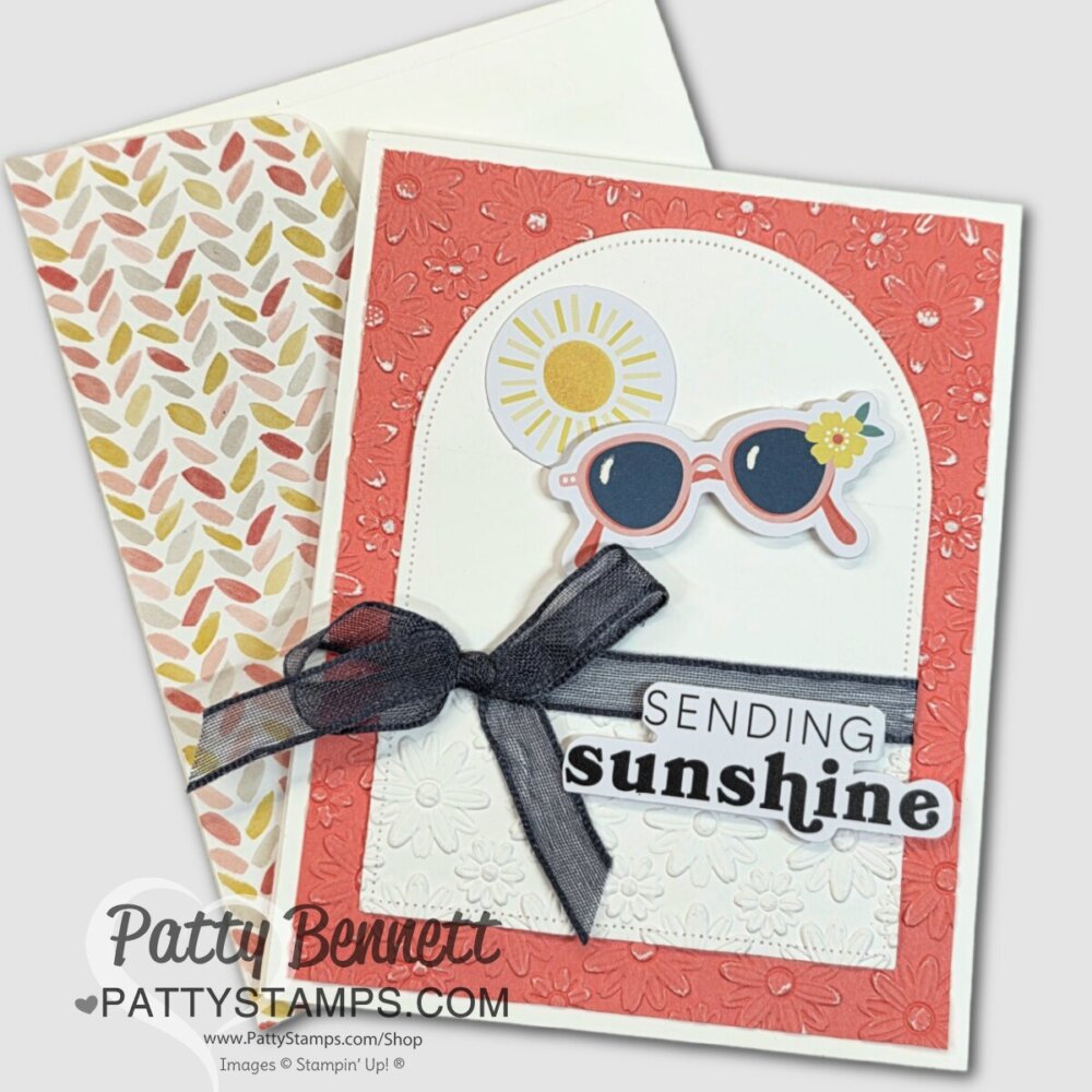 A handmade card features a textured coral border, a white floral-embossed panel, and a black ribbon bow. This quick & easy design is decorated with sun and sunglasses ephemera plus “SENDING sunshine” text. Leaf-pattern envelope beside it. To read more, visit Patty Stamps (pattystamps.com)