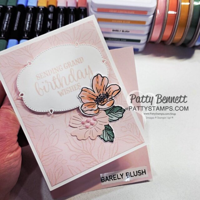 A hand holds a light pink floral birthday card with “Sending Grand Birthday Wishes” in gold on a white label. The card is decorated with orange-pink flowers and green leaves. In the background are colored markers, ink pads, and a “Barely Blush” label. To read more, visit Patty Stamps (pattystamps.com)