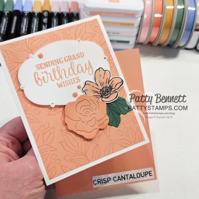 A hand holds a handmade birthday card featuring peach and cream colors, floral decorations, and the message "Sending Grand Birthday Wishes." Behind the card are markers and colored ink pads, with one labeled "Crisp Cantaloupe." The card is by Patty Bennett. To read more, visit Patty Stamps (pattystamps.com)