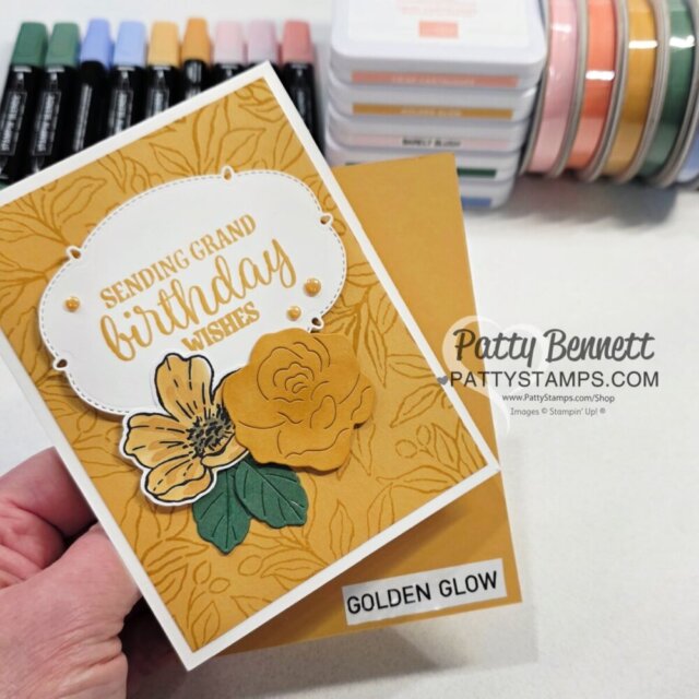 A hand holds a handmade card with gold floral designs, “Sending Grand Birthday Wishes” text, and yellow paper flowers. In the background are ink pads, colored markers, and craft supplies. The card base is labeled “GOLDEN GLOW.”. To read more, visit Patty Stamps (pattystamps.com)