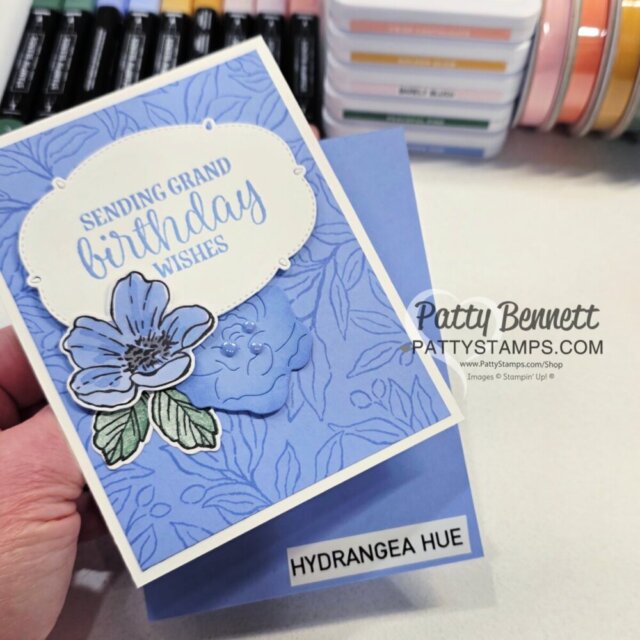 A hand holds a blue floral birthday card with a large flower and the message "Sending grand birthday wishes." Underneath is a matching blue envelope labeled "Hydrangea Hue." In the background are ink pads and markers stacked neatly on a desk. To read more, visit Patty Stamps (pattystamps.com)