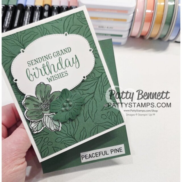 A hand holds a green floral handmade card with “Sending Grand Birthday Wishes” on a white label, decorated with embossed leaves, silver and green flowers. Below, a white label reads “PEACEFUL PINE.” Ink pads and markers are visible in the background. To read more, visit Patty Stamps (pattystamps.com)