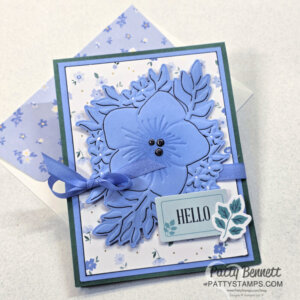 A handmade greeting card with a large blue layered flower and leafy die-cuts, blue ribbon bow, and “HELLO” sentiment on a tag. The background is white with a small blue floral pattern. A matching blue floral envelope is beneath the card. To read more, visit Patty Stamps (pattystamps.com)