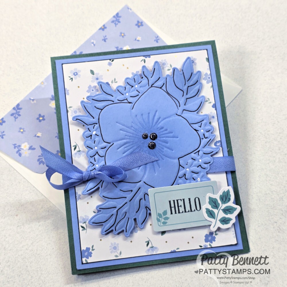 A handmade greeting card with a large blue layered flower and leafy die-cuts, blue ribbon bow, and “HELLO” sentiment on a tag. The background is white with a small blue floral pattern. A matching blue floral envelope is beneath the card. To read more, visit Patty Stamps (pattystamps.com)