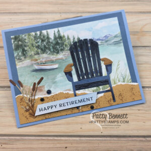 A handmade retirement card features a lake scene with trees, mountains, and a boat in the background. In the foreground, there's a die-cut Adirondack chair, cattails, sand, and a "HAPPY RETIREMENT" label. The card is signed "Patty Bennett. To read more, visit Patty Stamps (pattystamps.com)
