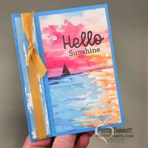 A handmade card features a colorful painted sunset over water with a small silhouette of a sailboat. The words “Hello Sunshine” appear in large black script and print. A yellow ribbon is tied vertically on the left, and a few white sequins decorate the scene. To read more, visit Patty Stamps (pattystamps.com)