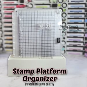 A clear stamp platform with a grid design stands upright in a white plastic holder. The background shows shelves filled with colorful crafting supplies. Text over the image reads, “Stamp Platform Organizer By StamptilDawn on Etsy.”. To read more, visit Patty Stamps (pattystamps.com)