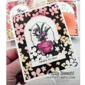 A hand holds a floral greeting card with a black, pink, and yellow flower-patterned background. The card features a pink vase with purple flowers on a white arch, a green gem embellishment, and a label reading “HERE’S TO YOU!” at the bottom. To read more, visit Patty Stamps (pattystamps.com)