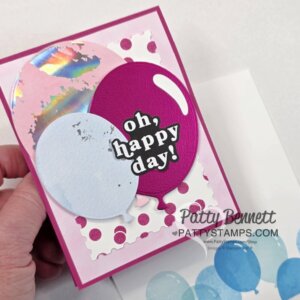 A hand holds a colorful greeting card with large pink and blue balloons, a shiny iridescent circle, and a white jagged frame. The card features the bold text “oh, happy day!” on a purple balloon. Below is the watermark: PattyBennett.com PattyStamps. To read more, visit Patty Stamps (pattystamps.com)