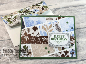 A handmade birthday card with a green border features patchwork-style panels of brown, blue, and beige floral patterns. “HAPPY BIRTHDAY to you” is stamped in green, accented by green dots. A matching floral envelope sits behind the card on a wooden background. To read more, visit Patty Stamps (pattystamps.com)
