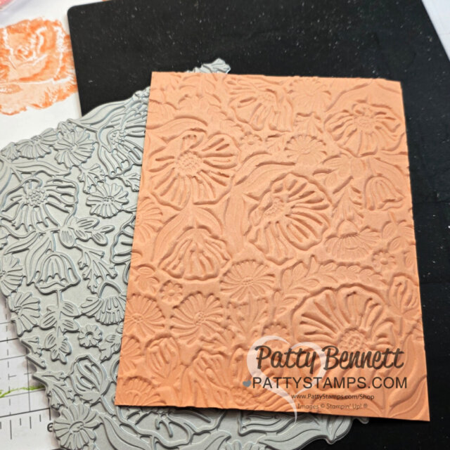 A peach-colored embossed paper with floral patterns lies atop a gray floral embossing folder. The background shows faint flower designs and a grid. The watermark reads "Patty Bennett PATTYSTAMPS.COM. To read more, visit Patty Stamps (pattystamps.com)
