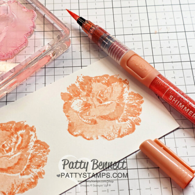 A peach-colored stamped rose on white paper, next to a matching brush pen and stamp block. The gridded work surface is visible. Patty Bennett’s watermark and website, pattystamps.com, appear at the bottom. The pen cap sits in the lower right corner. To read more, visit Patty Stamps (pattystamps.com)