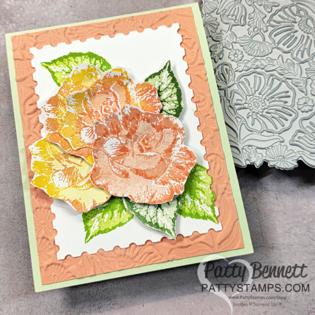 Handmade greeting card with layered, stamped roses in peach, yellow, and orange tones, surrounded by green leaves. The card has a peach-colored embossed frame on a white background and a light green border. A floral metal die and watermark "PattyStamps.com" are visible. To read more, visit Patty Stamps (pattystamps.com)