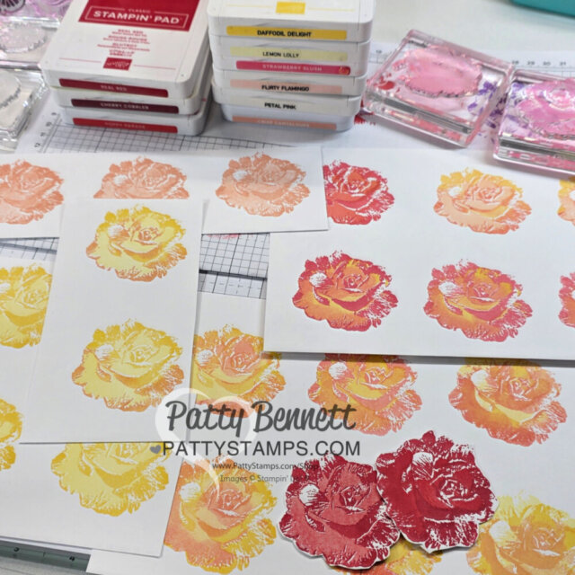 Hand-stamped rose images in pinks, yellows, and reds are displayed on white cardstock. Ink pads in various colors are stacked nearby. The workspace shows layered stamping techniques in progress. "Patty Bennett Pattystamps.com" watermark is visible. To read more, visit Patty Stamps (pattystamps.com)
